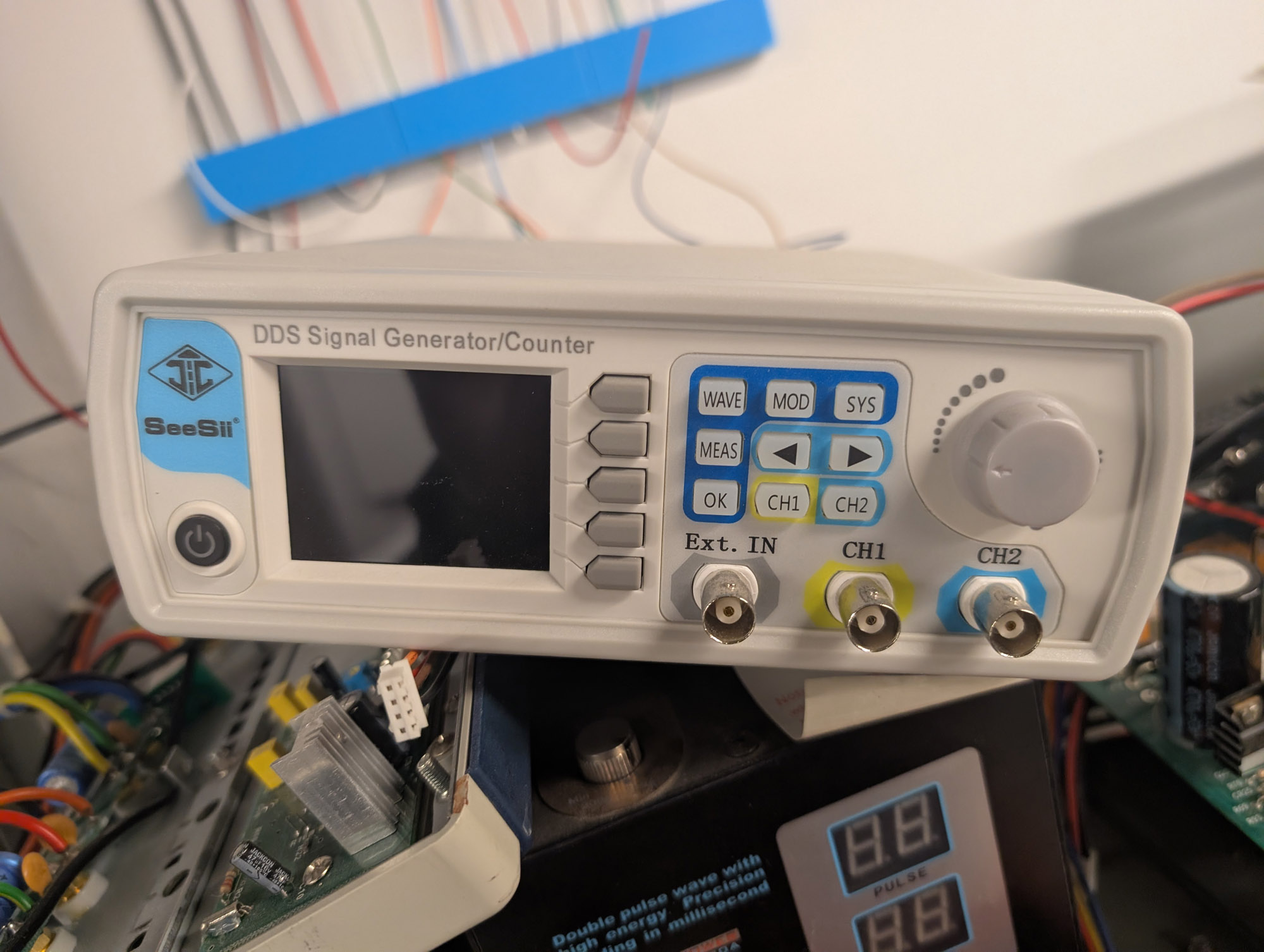 A beige plastic instrument with SeeSii written on the left above a round power button, a small LCD with "DDS Signal Generator/Counter" beside that, and 5 softkeys beside the screen. A small array of 9 buttons and an unlabelled knob are beside that, and BNC inputs for two channels plus Ext In at the bottom right.